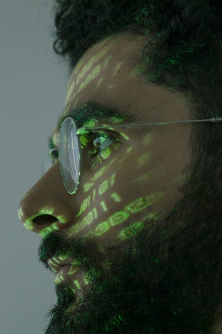 A close-up profile of a man with dark, curly hair and facial hair, wearing round glasses, with a projection of green digital code and patterns on his face and glasses. The background is neutral gray, 