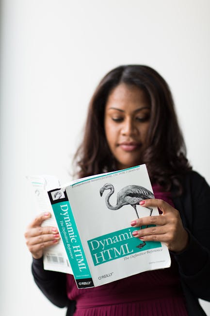 The image features a woman with dark, wavy hair reading a book titled 'Dynamic HTML'. She is wearing a black blazer over a deep burgundy top and appears focused on her reading. The background is plain